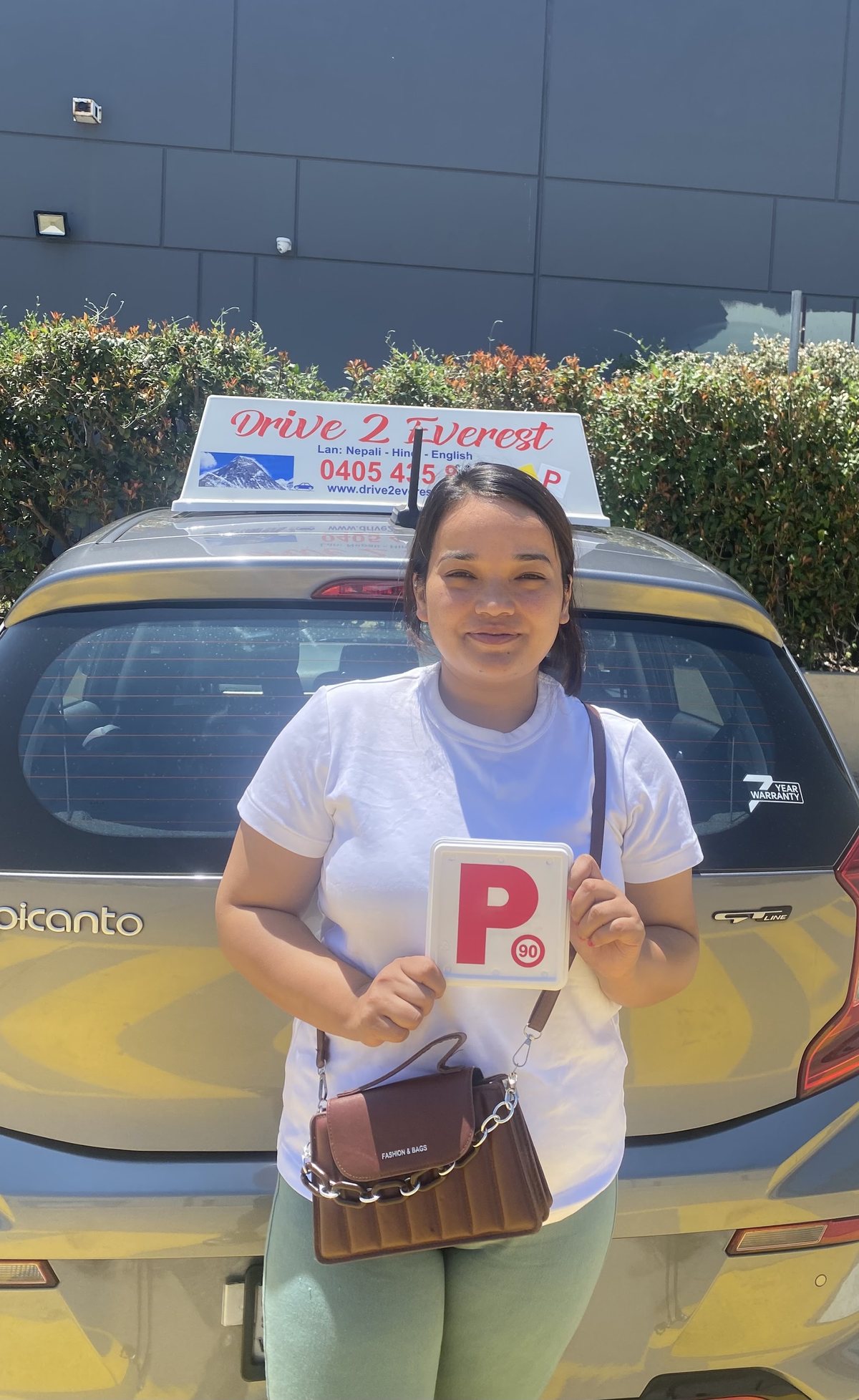 A new driver holding a red P-plate in front of the Drive 2 Everest car