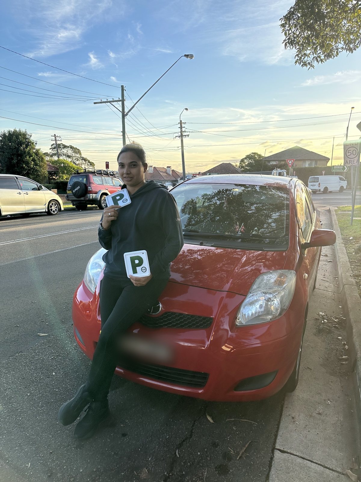 A new driver sitting on a red car holding two green P-plates at sunset